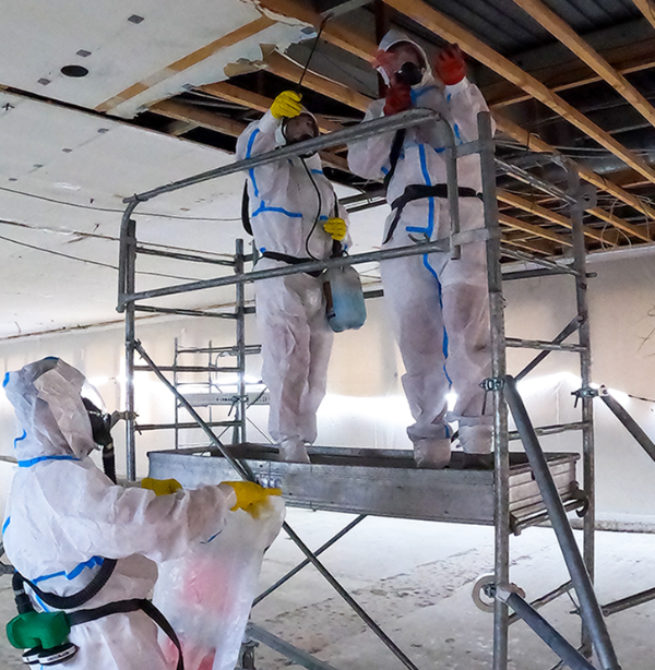 Asbestwerkers in beschermpakken op steigers voeren sanering uit in een afgesloten binnenruimte.